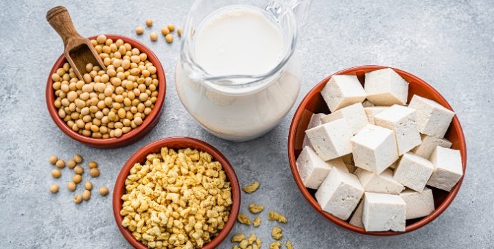 Eine Karaffe mit Sojamilch, und Schalen mit Tofuwürfeln, Sojabohnen und texturiertem Sojaprotein stehen auf einer hellen Tischplatte