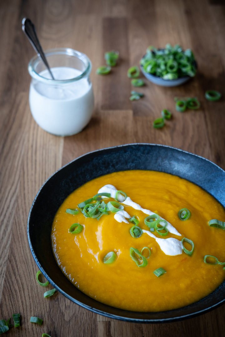 Ein Teller Kürbis-Kokos-Cremesuppe, ein Schälchen mit Frühlingszwiebeln und ein Glas mit Kokossahne