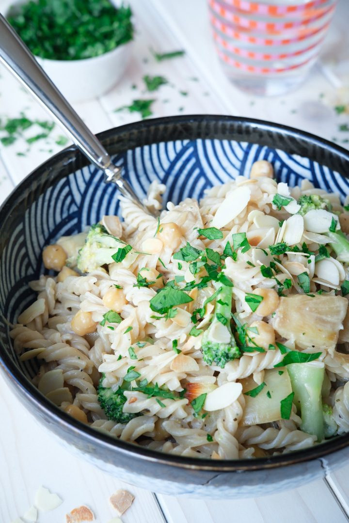 Ein Teller vegane Pasta mit karamellisierten Zwiebeln, Kichererbsen und Brokkoli, getoppt mit frischer Petersilie, steht auf einem weißen Holztisch