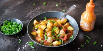 Ein Teller mit veganen Patatas bravas steht auf einer dunklen Tischplatte, daneben ein Schälchen mit Schnittlauch und eine Flasche mit scharfer Würzsauce.