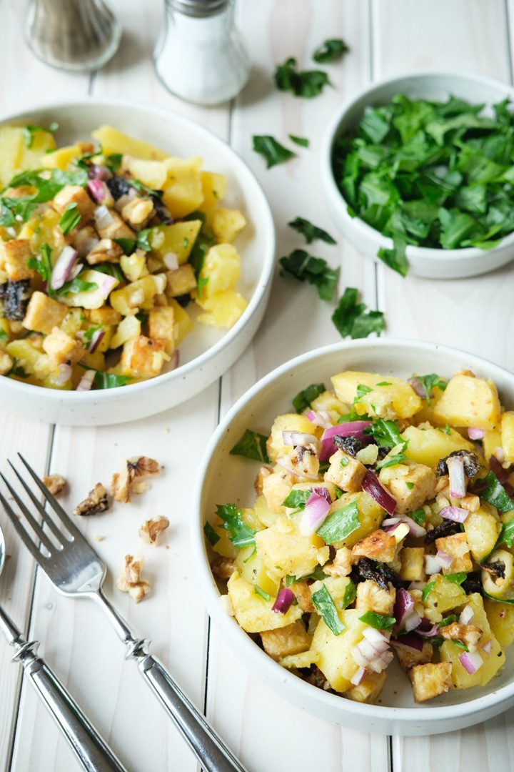 Zwei Teller mit veganem Kartoffelsalat stehen auf einer weißen Tischplatte aus Holz, daneben steht ein Schälchen mit gehackter Petersilie sowie ein Salz- und Pfefferstreuer