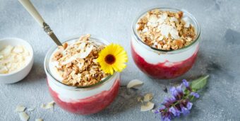 Zwei Gläser mit Rhabarber-Himbeer-Kompott, Kokossahne und Knuspermüsli stehen auf einer hellen Tischplatte