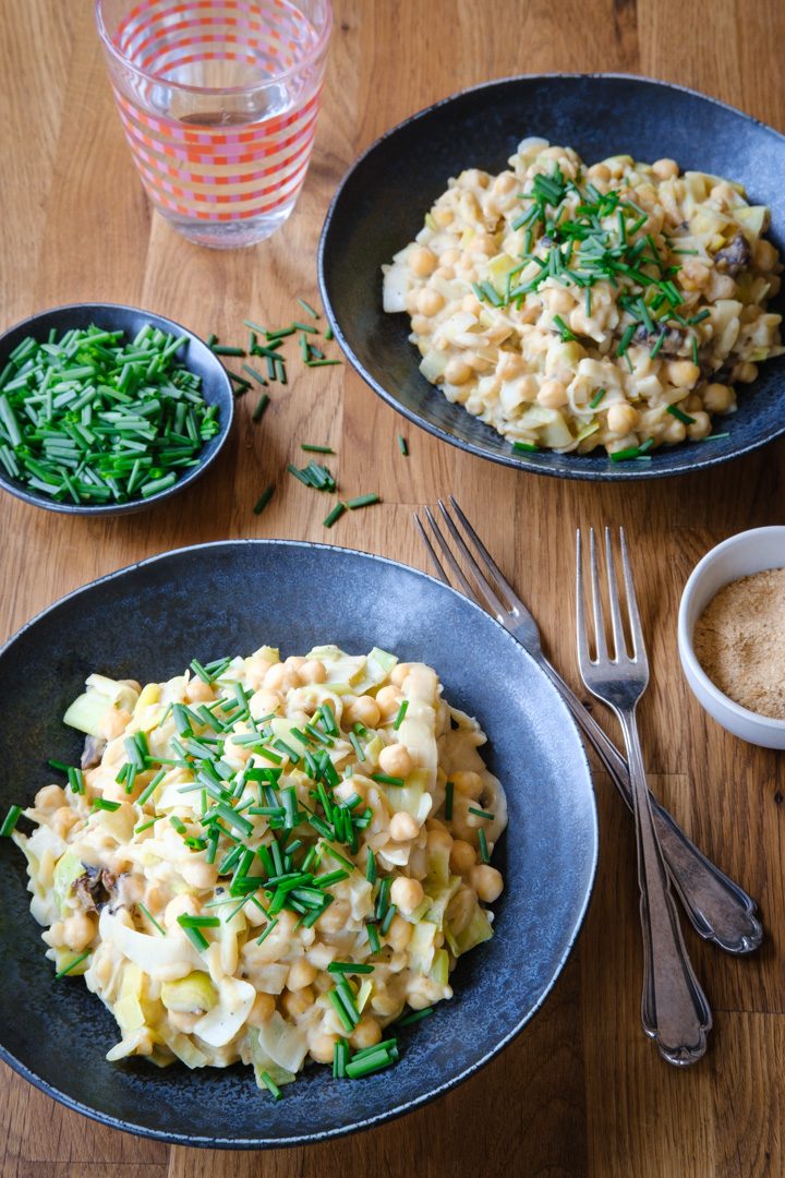 Zwei dunkle Teller mit der Lauch-Kichererbsen-Pfanne und einem Schälchen mit frischem Schnittlauch. Daneben steht ein Wasserglas