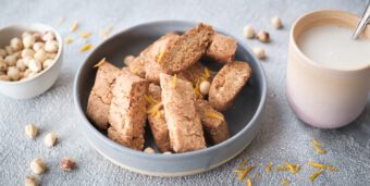 Vegane Cantuccini liegen in einer Schale, daneben steht eine Tasse mit Milch und ein Schälchen mit Haselnüssen