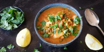 Eine Schale veganes Linsen-Blumenkohlcurry steht auf einer dunklen Tischplatte mit Zitronen und verstreutem Koriander