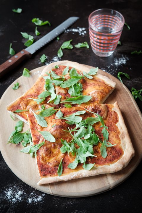 Zwei Stücke vegane Pizza Marinara mit Rucola liegen auf einem Holzbrettchen