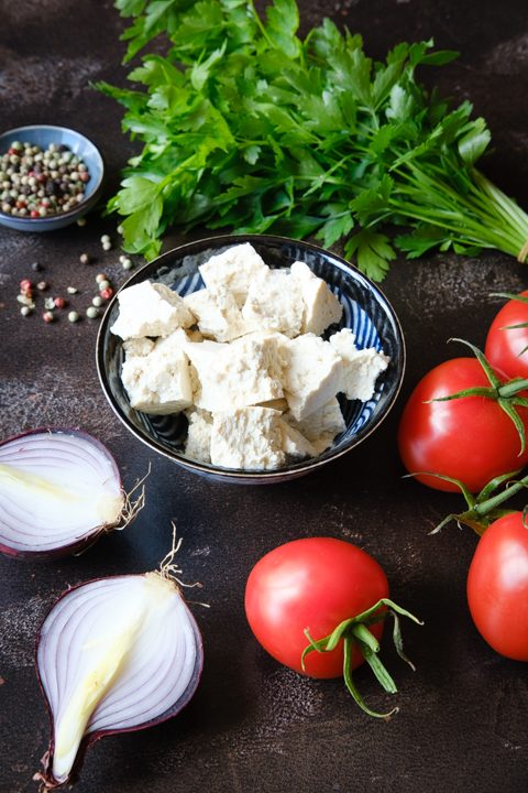 Naturtofu, ein Bund Petersilie, zwei Zwiebelhälften und Tomaten liegen auf einer dunklen Tischplatte