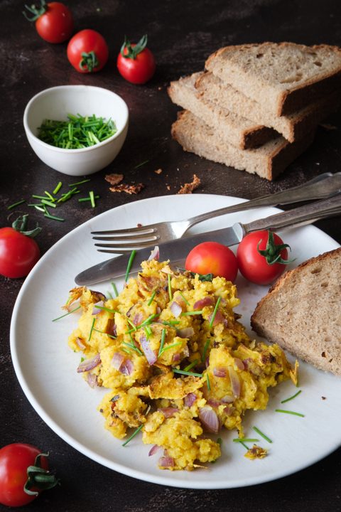 Veganes Rührei aus Mung Dal mit roten Zwiebeln und Schnittlauch auf einem weißen Teller, dazu Tomaten und Brot.