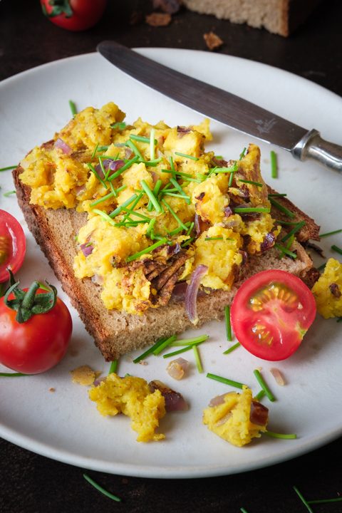 Veganes Mung-Dal-Rührei auf einer Scheibe Brot, garniert mit Schnittlauch und roten Zwiebeln, dazu Tomaten auf einem weißen Teller.