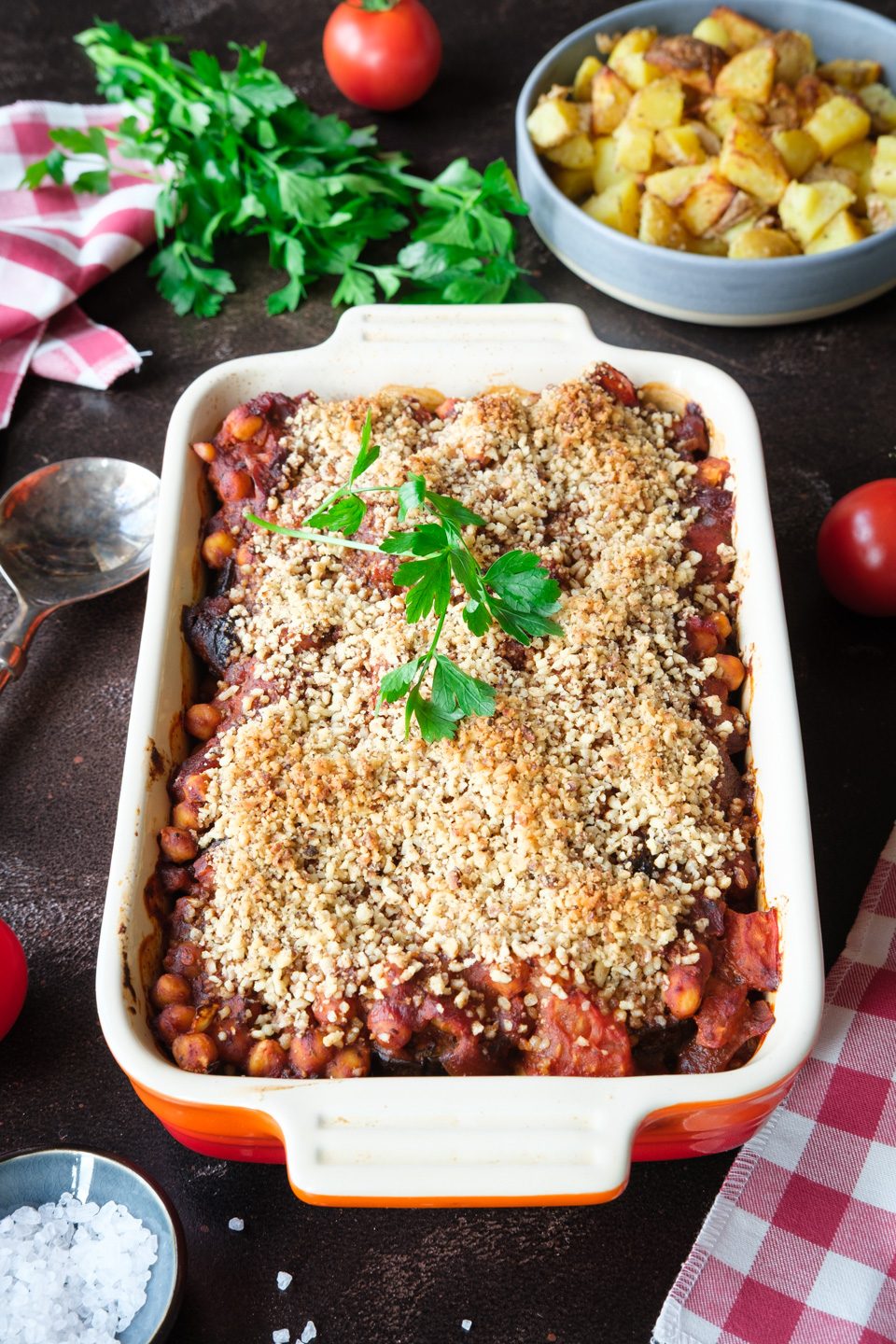 Frisch gebackener Auberginenauflauf mit Kichererbsen und goldbraunem Haselnuss-Topping, daneben eine Schüssel mit Kartoffelwürfeln