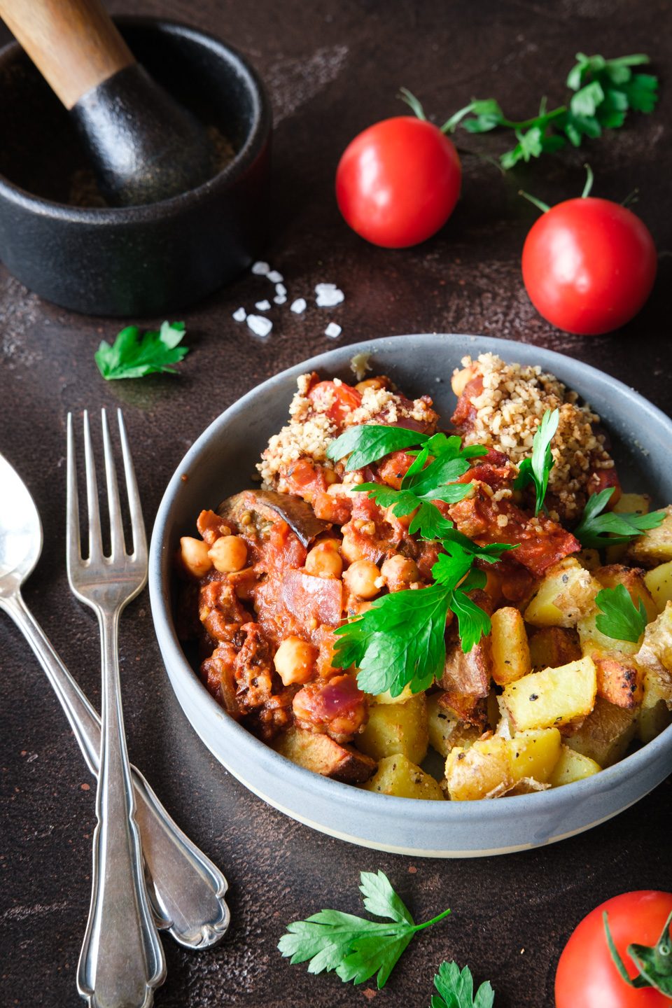 Portion Auberginen-Kichererbsen-Auflauf mit knusprigen Kartoffelwürfeln und frischer Petersilie in einer grauen Schüssel